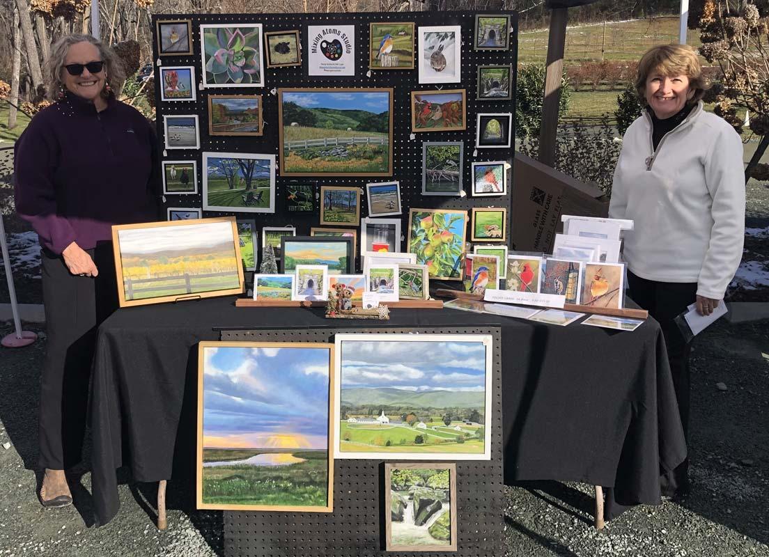 Penny and Debbie smiling in front of a display table of their artwork at a local art festival